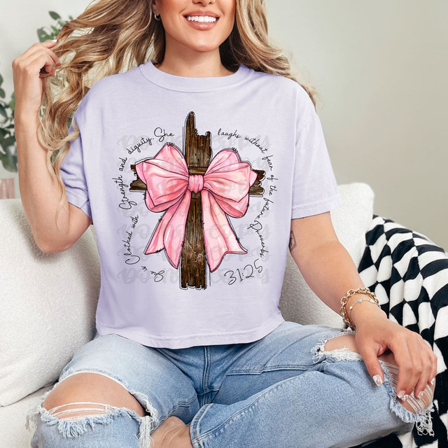 She Is Clothed With Strength And Dignity Bow Cross DTF Heat Transfer, image of a woman wearing a white T-shirt with a decorative cross and pink bow design, featuring motivational script around the design.