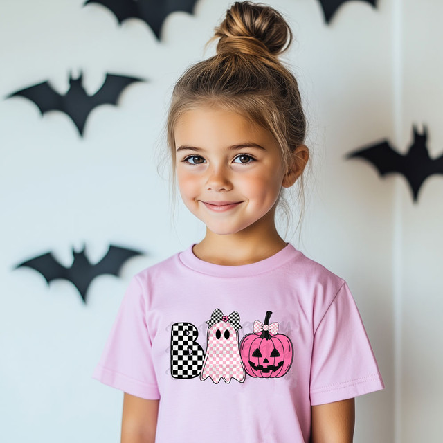 Boo Pink Checkered Ghost DTF Heat Transfer, product image of a girl wearing a pink T-shirt with Halloween ghost and pumpkin designs, Halloween decorations in the background.
