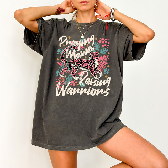Praying Mama Raising Warriors DTF Heat Transfer, a woman wearing an oversized black t-shirt with colorful graphic design of a pink leopard and tropical leaves, showcasing the product.