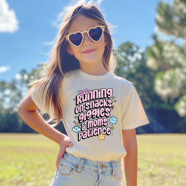 Running On Snacks, Giggles & Moms Patience DTF Heat Transfer on a white T-shirt worn by a young girl outside in a park with sunglasses.