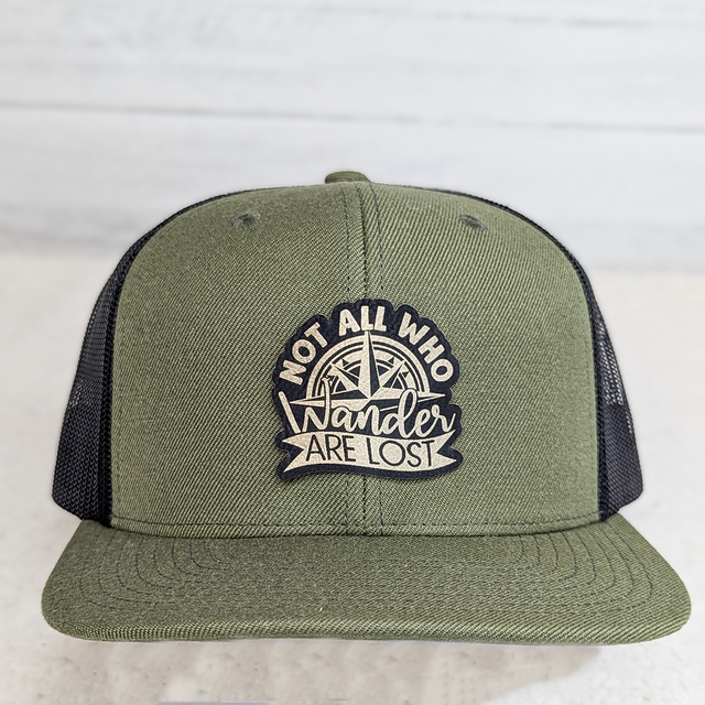 Not All Who Wander Are Lost FAUX Leather Patch, green and black trucker cap with embroidered patch featuring a compass and the phrase 'Not All Who Wander Are Lost'.