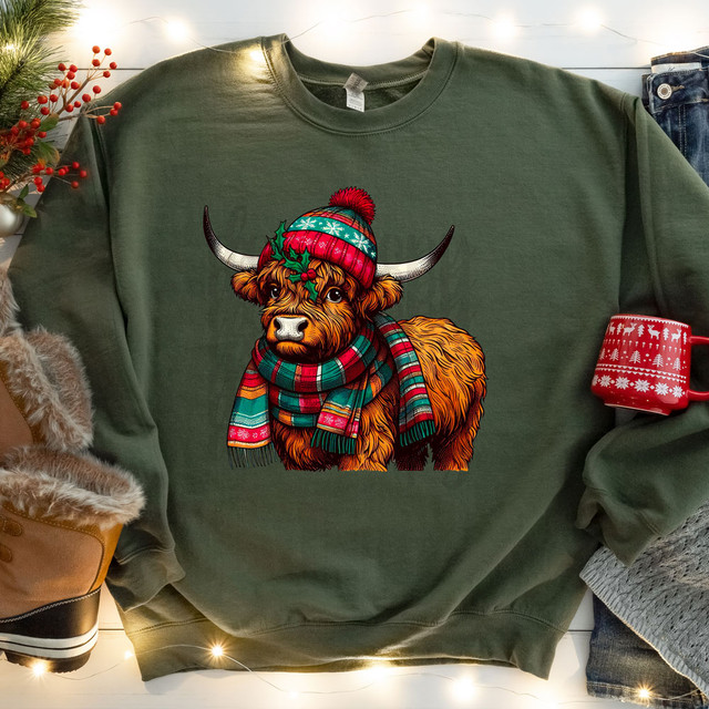Christmas Highland Cow DTF Heat Transfer, a colorful illustration of a Highland cow wearing a festive red and green winter hat and plaid scarf on a green sweatshirt.