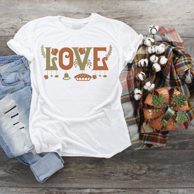 Love Thanksgiving Sublimation Transfer, white T-shirt with Thanksgiving-themed design, including the word LOVE and decorative elements, displayed on a rustic wooden surface with jeans, plaid blanket, and cotton flowers.