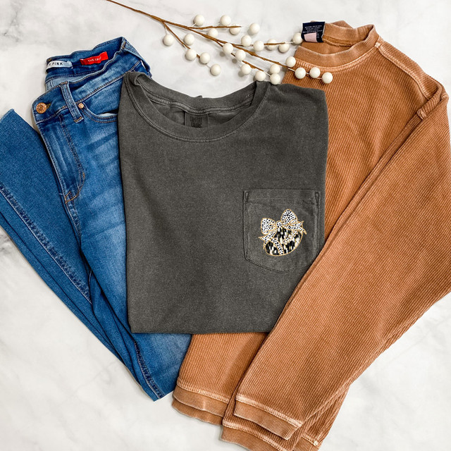 Black/White Brushstroke Football Coquette Faux Glitter DTF Heat Transfer, image showing a gray T-shirt with a pocket detail, blue jeans, and a brown ribbed sweater on a white background with decorative white berry branch.