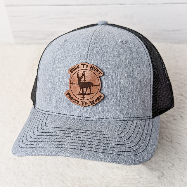 Born To Hunt Forced To Work Leather Hat Patch, embroidered patch with a deer silhouette and crosshairs design on a grey and black trucker hat.