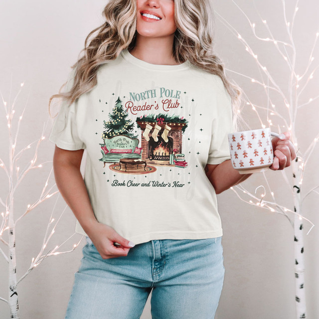 North Pole Reader's Club DTF Heat Transfer, festive Christmas print on a T-shirt featuring a decorated fireplace, Christmas stockings, and a Christmas tree.