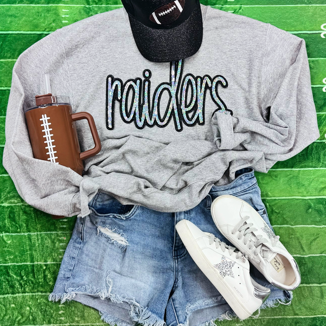 Raiders Sequin Patch, a stylish grey sweatshirt with a sparkling 'raiders' sequin patch, paired with ripped denim shorts, white sneakers with glitter accents, a football-themed cap, and a brown football-themed water bottle, laid out on a vibrant green surface.