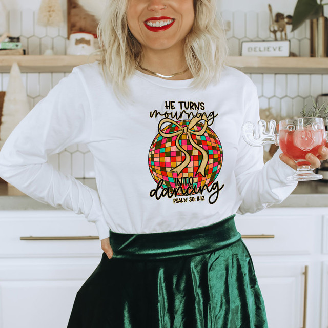 He Turns Mourning Into Dancing DTF Heat Transfer, woman wearing a white shirt with colorful graphic and inspirational quote, holding a pink pig-shaped glass, in a modern kitchen.