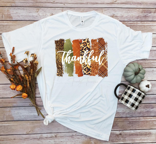 Thankful word art fall colors plaid leopard glitter pattern sublimation transfer, on a white T-shirt with fall-themed decorations around, including dried flowers and pumpkins.