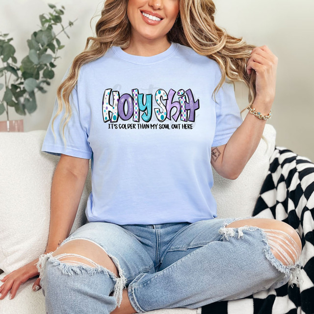 Holy Shit It's Colder Than My Soul In Here DTF Heat Transfer T-Shirt, a woman wearing a light blue t-shirt with bold, colorful text and humorous phrase, sitting on a couch with a relaxed posture.