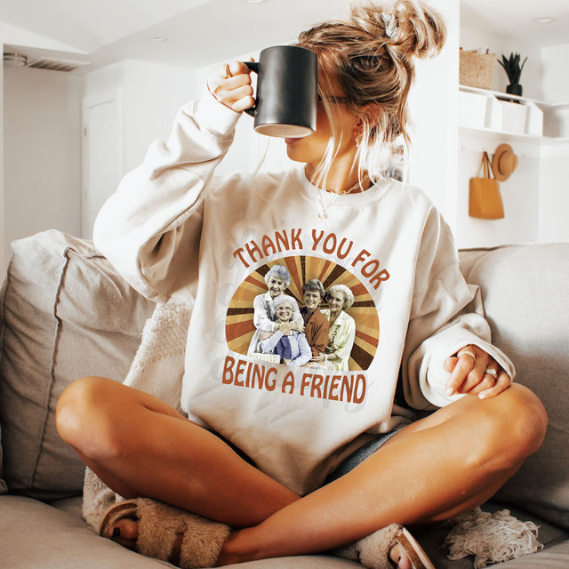Retro Thank You For Being A Friend DTF Heat Transfer, a woman sitting cross-legged on a couch wearing a sweatshirt with a vintage design of elderly friends and the message 'Thank You For Being A Friend' in a cozy home setting.