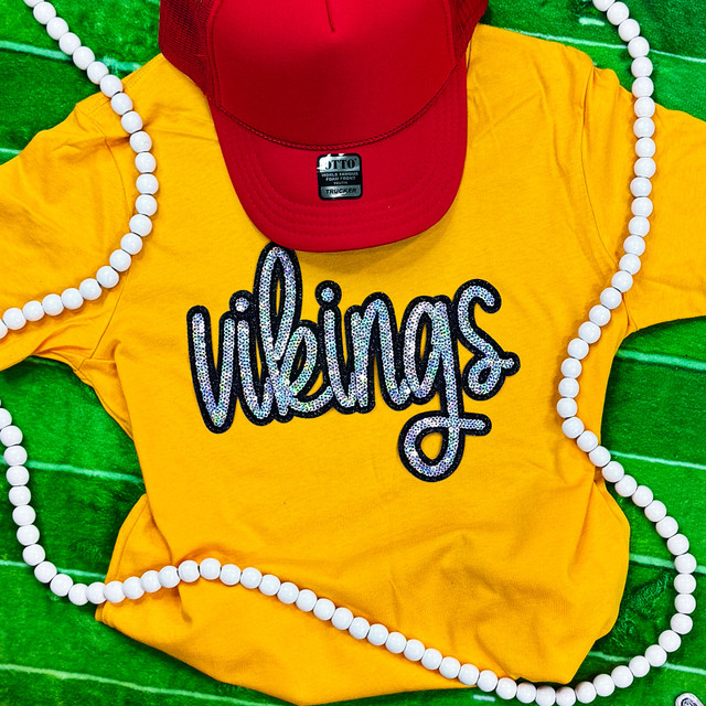 Vikings YOUTH Sequin Patch, yellow T-shirt with the word 'vikings' decorated with colorful sequins and rhinestones, red cap, and white bead necklace, on green textured background.