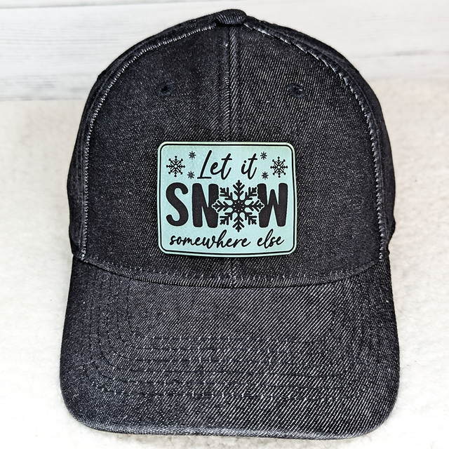 Let It Snow Somewhere Else FAUX Leather Patch, black baseball cap with a decorative patch featuring snowflakes and the phrase "Let it Snow somewhere else" in black text.