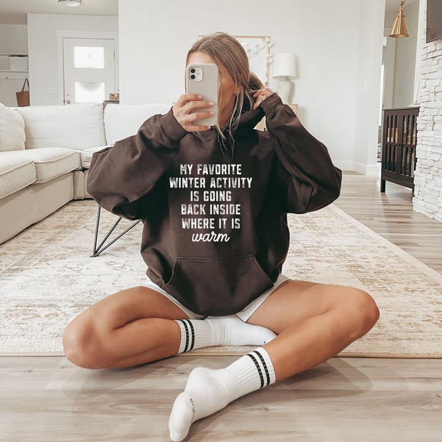 My Favorite Winter Activity In Going Back Inside Where It Is Warm WHITE DTF Heat Transfer, a woman wearing a black hoodie with this printed phrase sitting on the floor in a cozy living room.