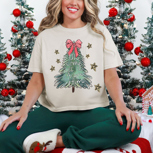 Oh Come Let Us Adore Him Hand Drawn Screen Print Heat Transfer, Christmas themed product featuring a woman sitting in front of decorated Christmas trees, wearing a cream-colored T-shirt with a festive green tree illustration and the phrase 'Oh Come Let Us Adore Him' with a pink bow at the top.