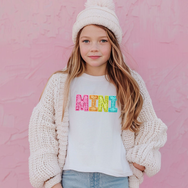 Bright Groovy Mini Faux Sequins DTF Heat Transfer, colorful mini sequin letters spelling 'MNI' on a white t-shirt worn by a young girl with a pink background.