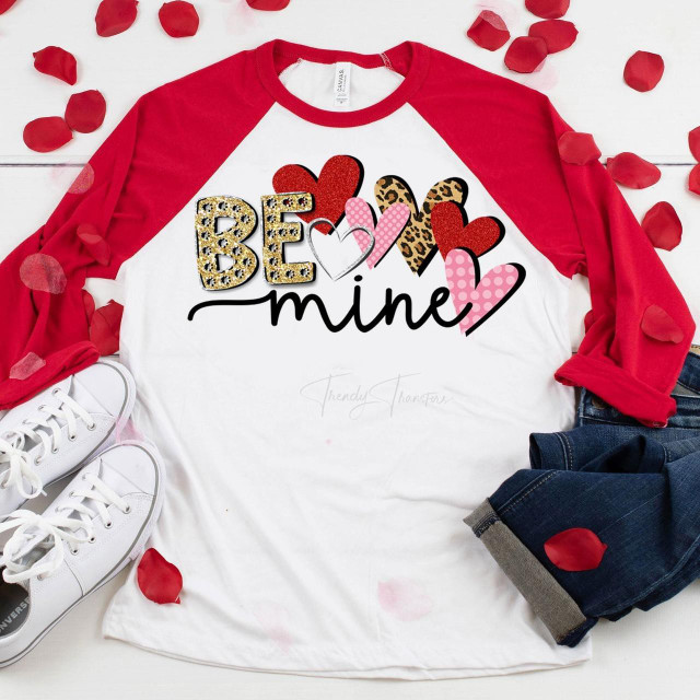 Be Mine Glitter Valentine Sublimation Transfer, a colorful design with hearts and the words 'Be Mine' on a white and red raglan shirt, surrounded by red rose petals, white sneakers, and folded jeans.
