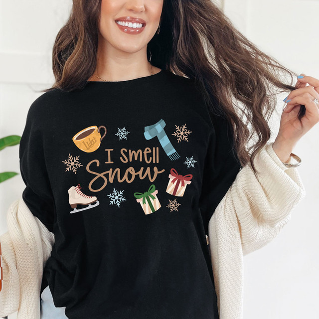 I Smell Snow DTF Heat Transfer, detailed graphic design on a black T-shirt featuring a coffee mug, ice skate, gift boxes, snowflakes, and a scarf with the text 'I Smell Snow'.