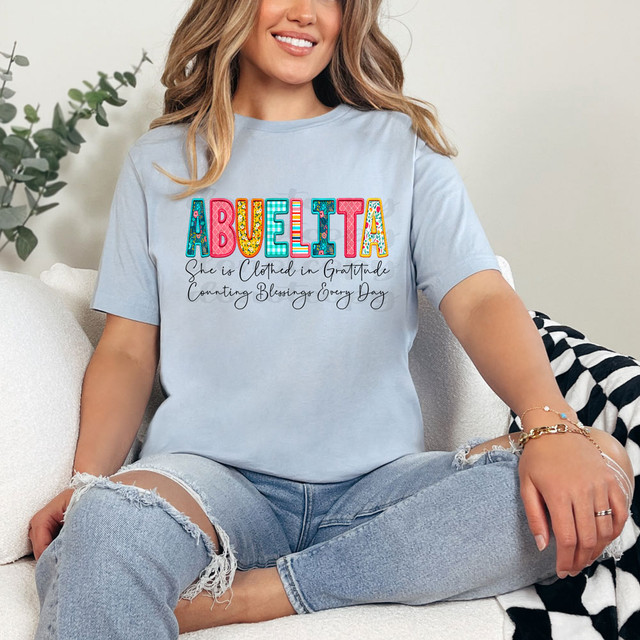 Abuelita She Is Clothed In Gratitude Counting Blessings Every Day DTF Heat Transfer, woman wearing a light blue T-shirt with colorful text and a heartfelt message, sitting on a cozy sofa with a white pillow and a plant in the background.