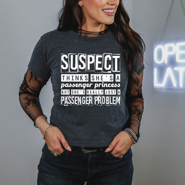 Suspect Thinks She's A Passenger Princess But She's Really Just A Passenger Problem DTF Heat Transfer, a woman wearing a dark gray t-shirt with bold white and black text, smiling in a casual setting.