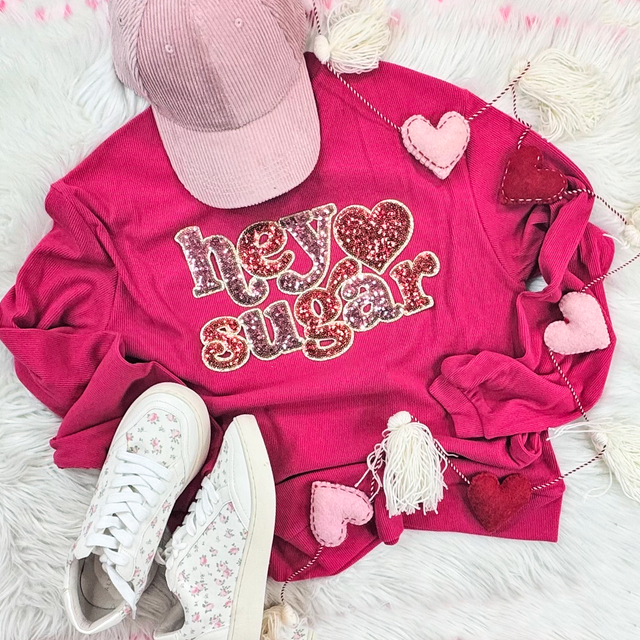 Hey Sugar Sequin Patch, pink sweatshirt with sparkly sequined lettering and heart details, casual shoes, and decorative hearts on a soft furry background.