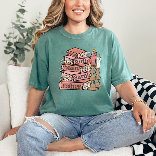 Women Of The Bible Books DTF Heat Transfer, a woman wearing a green T-shirt with a graphic design of stacked books labeled with biblical names and candles.