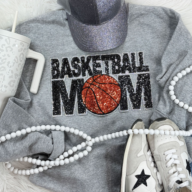 Basketball Mom Sequin Patch, close-up of a gray sweatshirt with a shiny, glittery sequin patch of a basketball and the words 'Basketball Mom', surrounded by white sneakers, a pearl necklace, a white mug, and a sparkly cap.