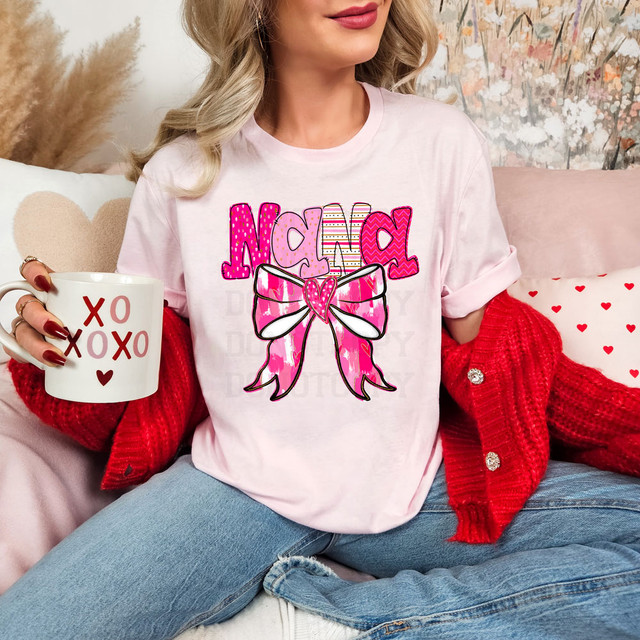 Nana Valentine Coquette DTF Heat Transfer, pink graphic T-shirt featuring a large bow with a heart, worn by a smiling woman sitting on a cozy couch with Valentine's Day themed pillows.