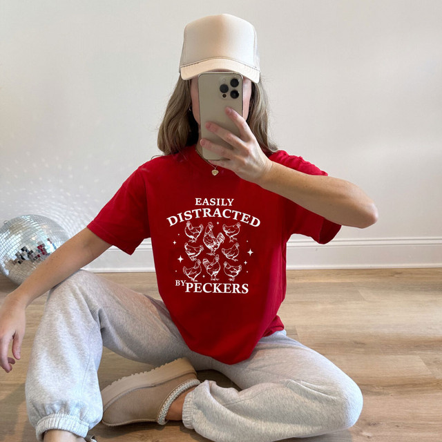 Easily Distracted By Peckers WHITE DTF Heat Transfer, white text and chicken graphics on a red T-shirt worn by a person sitting on the floor.