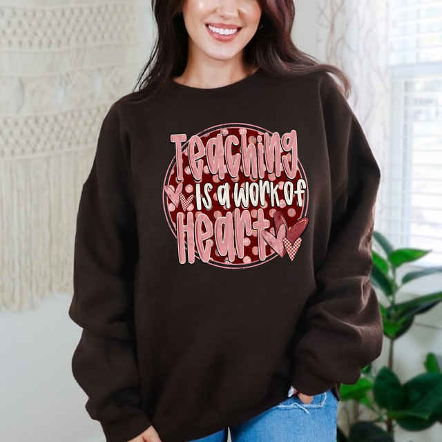 Teaching Is A Work Of Heart Polka Dots DTF Heat Transfer, a close-up of a woman wearing a brown sweatshirt with a pink and white heart-themed graphic and the phrase 'Teaching Is A Work Of Heart'.