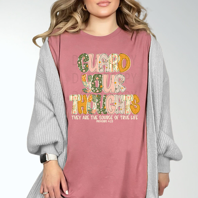 Guard Your Thoughts They Are The Source Of True Life CREAM DTF Heat Transfer, colorful printed design on a pink t-shirt worn by a woman with a gray cardigan.
