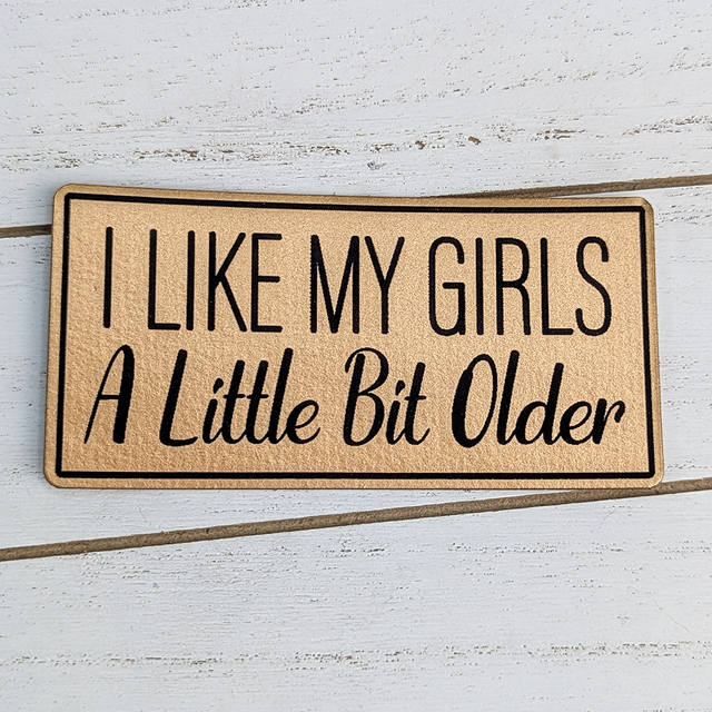 Product: I Like My Girls A Little Bit Older Faux Leather Patch, a beige furry patch with black embroidered text and a black border, placed on a white wooden surface.