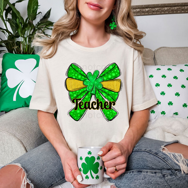 Teacher St Patrick's Day Coquette Bow DTF Heat Transfer, a green and yellow bow design with the word Teacher, worn by a woman holding a shamrock-themed mug