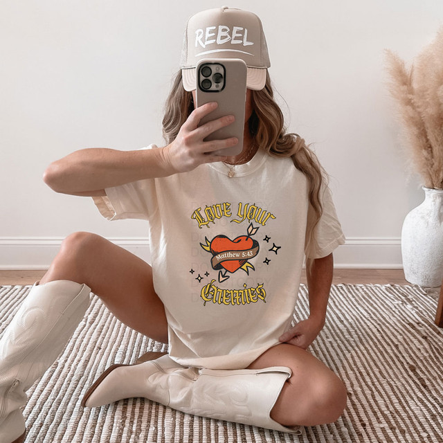 Love Your Enemies Red Heart DTF Heat Transfer, woman wearing graphic t-shirt with heart and Bible verse, sitting on rug with beige boots, beige cap, and neutral decor.