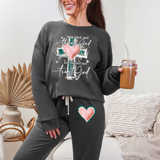 Look Back And Thank God Look Forward And Trust God White Set DTF Heat Transfer, image of a woman wearing a matching sweatshirt and sweatpants with spiritual text and heart graphics, holding iced coffee with a straw.