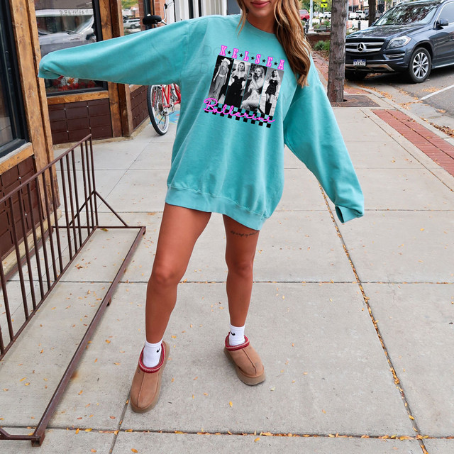 KB Collage DTF Heat Transfer, a woman wearing a turquoise oversized sweatshirt with a black and white graphic print and pink text, standing on a sidewalk in an urban setting.