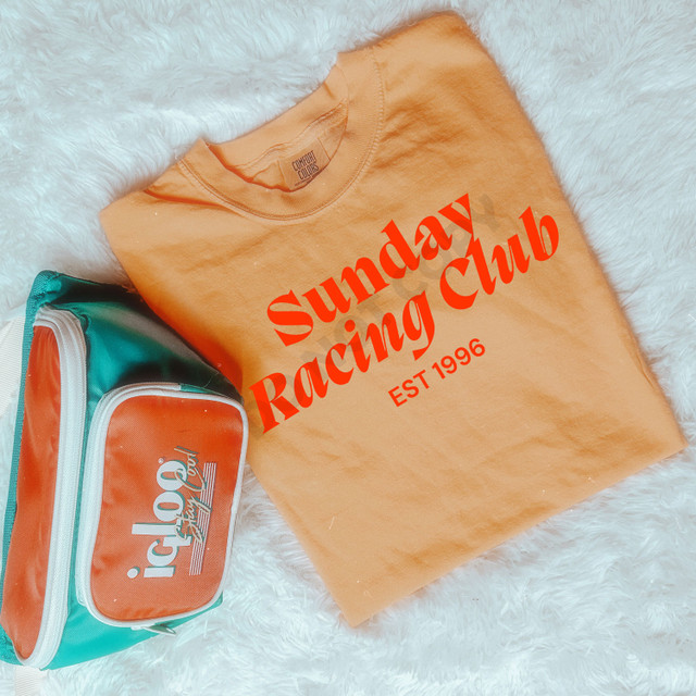 Sunday Racing Club DTF Heat Transfer, orange T-shirt with red printed logo and text on a soft white furry surface.