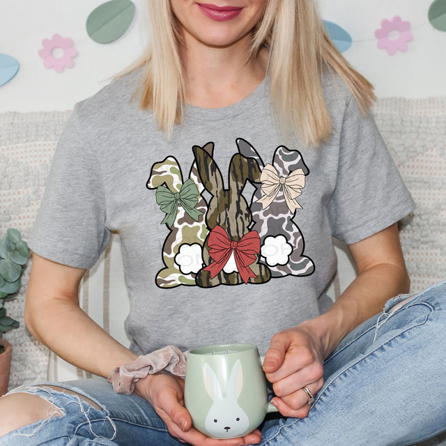 Camo Patterned Bunnies DTF Heat Transfer, a graphic design featuring three camo patterned rabbits with bows on their ears, printed on a gray T-shirt worn by a woman.