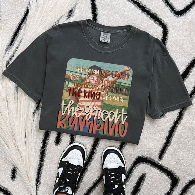 The Great Bambino DTF Heat Transfer, a colorful graphic t-shirt with football-themed text and images, laid flat on a patterned rug with sneakers below.