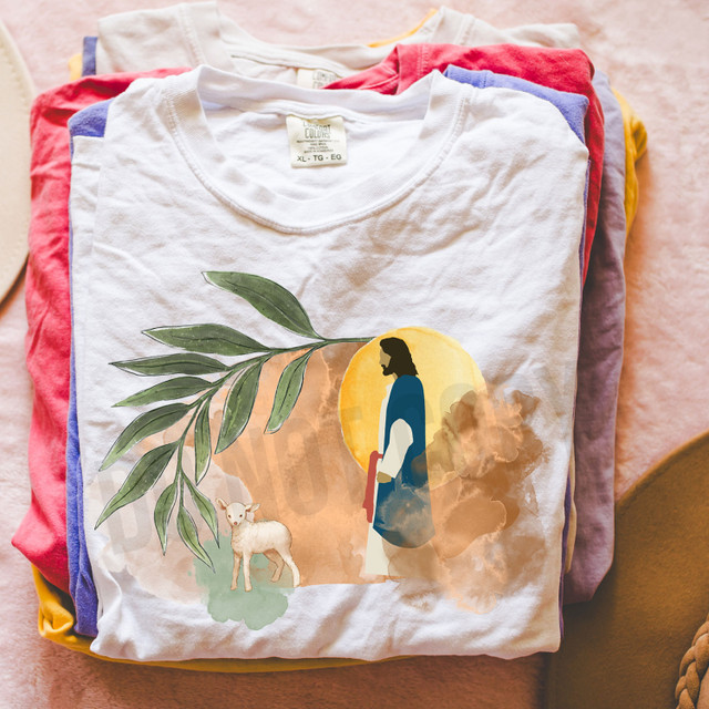 Watercolor Jesus DTF Heat Transfer, colorful T-shirt with artistic depiction of Jesus, a lamb, green leaves, and a sun in the background.