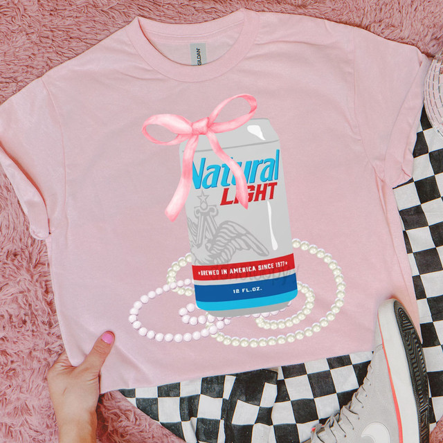 Natty Lt Coquette Bow DTF Heat Transfer, pink t-shirt with a print of a can of Natural Light beer decorated with a pink bow, pearl necklace design, and a hand holding the shirt, on a pink furry surface with black and white checkered cloth and a white sneaker.