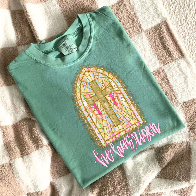 He Has Risen Stained Glass DTF Heat Transfer on a mint green T-shirt, featuring a colorful stained glass design with a cross and the word 'Resurrection' in pink cursive font.
