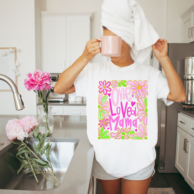 One Loved Mama Faux Glitter DTF Heat Transfer, image of a woman with towel on head drinking from a pink mug in a kitchen, wearing a white t-shirt with colorful floral and text design.