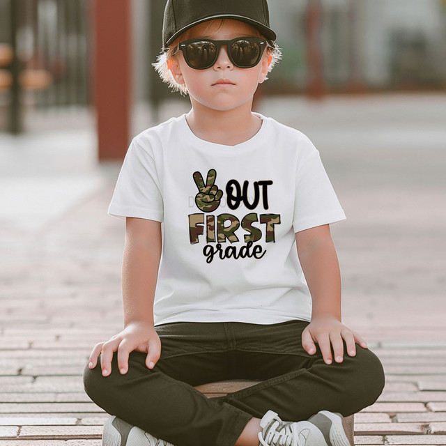 Camo Peace Out First Grade DTF Heat Transfer, a young boy wearing a white t-shirt with a camo peace sign hand gesture and camo letters for 'First', sitting outdoors.