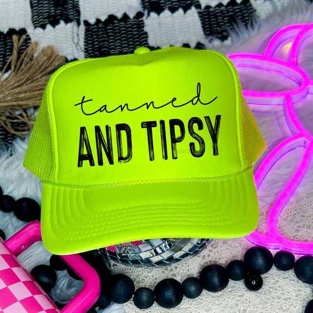Minimalist Tanned And Tipsy BLACK DTF Heat Transfer, neon yellow trucker cap with black text, surrounded by colorful accessories and decor.