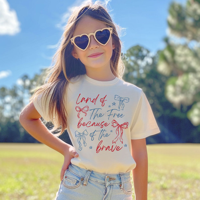 Land Of The Free Because Of The Brave Coquette Bows DTF Heat Transfer, a young girl wearing sunglasses and a graphic T-shirt with patriotic bows and stars printed on it outdoors.