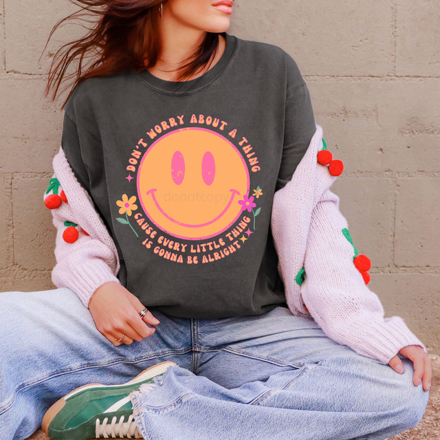 Don't Worry About A Thing Retro Happy Face DTF Heat Transfer, a woman wearing a dark grey sweatshirt with a pink smiling face graphic and a motivational quote, paired with light jeans and green sneakers, sitting against a beige brick wall.