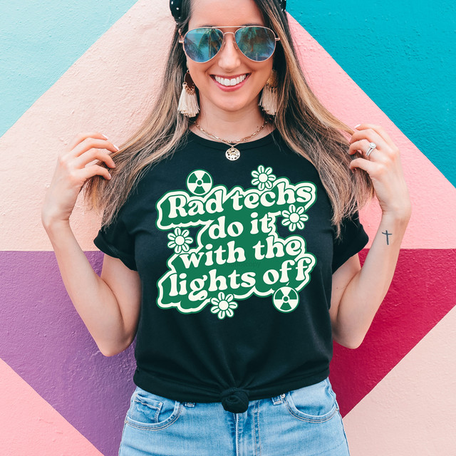 Rad Techs Do It With The Lights Off DTF Heat Transfer, a woman wearing a black t-shirt displaying the slogan and graphics, standing against a colorful geometric wall.