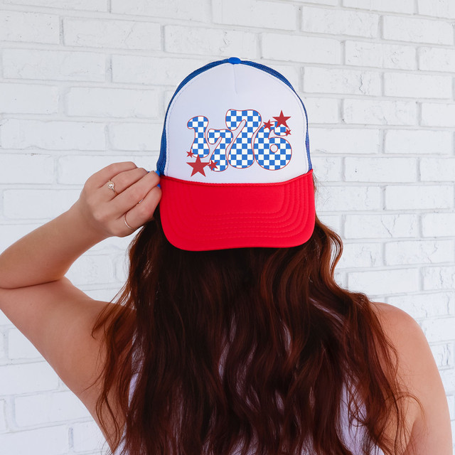 Retro 1776 DTF Heat Transfer, colorful patriotic design on a cap with a red brim, blue and white checkered number 1776 with stars, worn by a woman with long brown hair against a white brick wall.