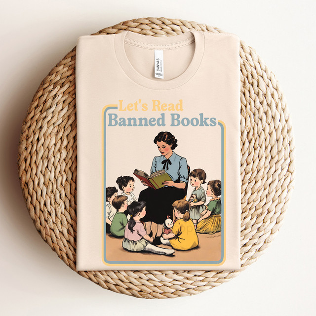 Let's Read Banned Books DTF Heat Transfer, a vintage-style illustration of a woman reading a book to children, on a folded cream-colored T-shirt placed on a woven round placemat.